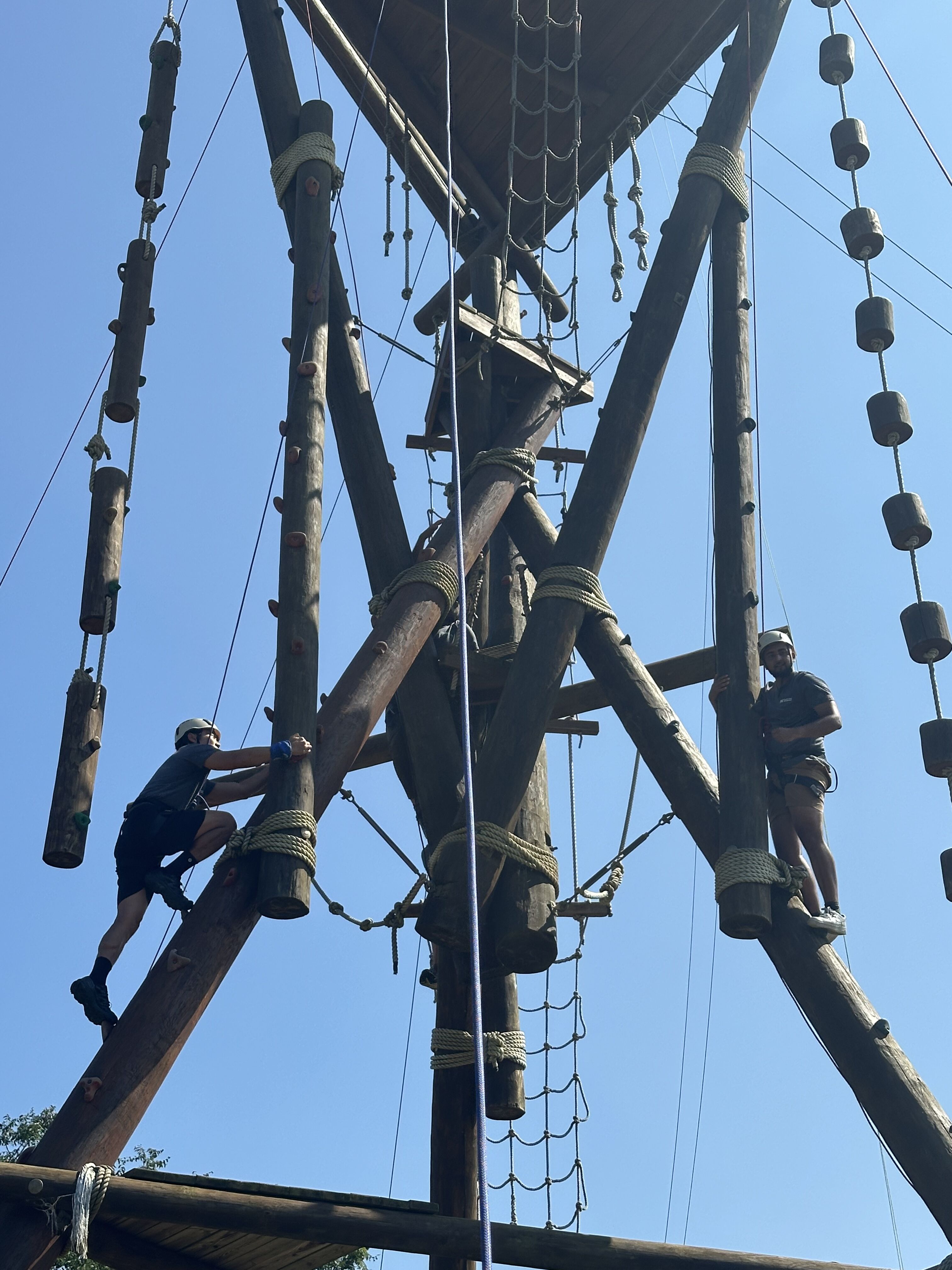 This image captures a thrilling adventure course with two individuals actively participating. One person is seen climbing a wooden structure, while another stands on a platform, both equipped with safety gear. The obstacle course appears to be set against a clear blue sky, suggesting a perfect day for outdoor activities. The intricate wooden beams and ropes create a challenging and exciting environment for participants.